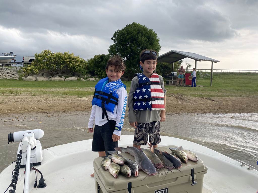 A cute photo of two kids wearing life vests while standing on a charter fishing boat docked near the shore and posing behind a cooler with fish caught that day during a Red River angling trip