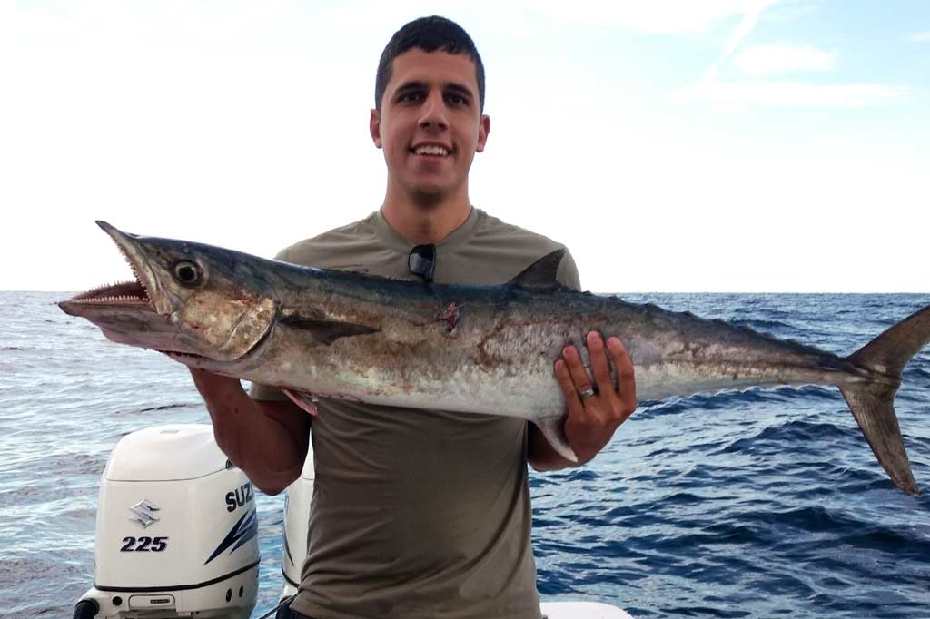 A smiling young man holding a big King Mackerel, at the back of a boat, with open waters in the background