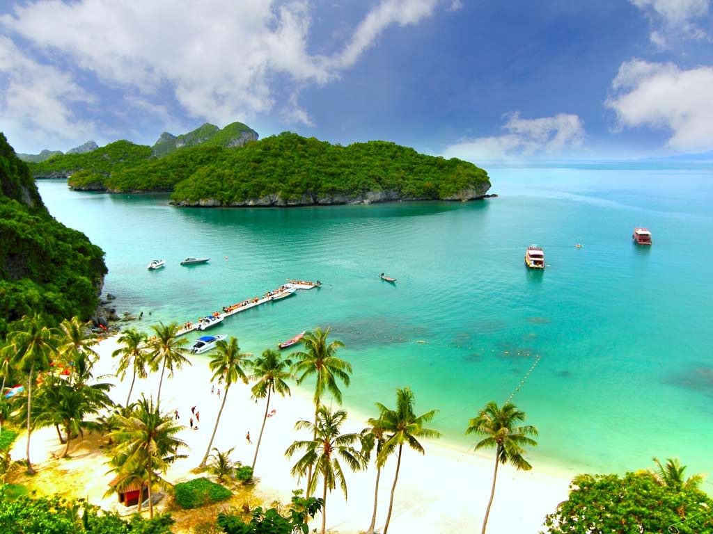 An aerial shot of a beach in Koh Samui with palm trees and sand visible in the forefront and azure waters, fishing boats, and another island visible in the background on a cloudy day.