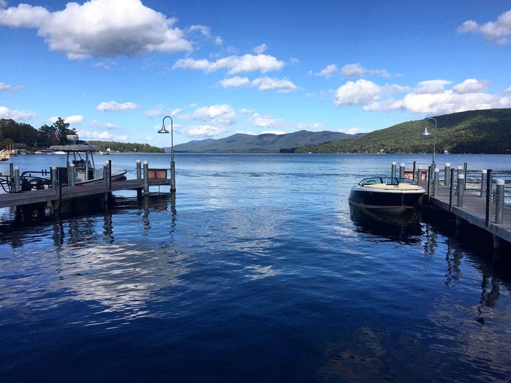 A view from the shore towards the waters of Lake George with two docks on either side of the image and a boat moored against each on a sunny day.