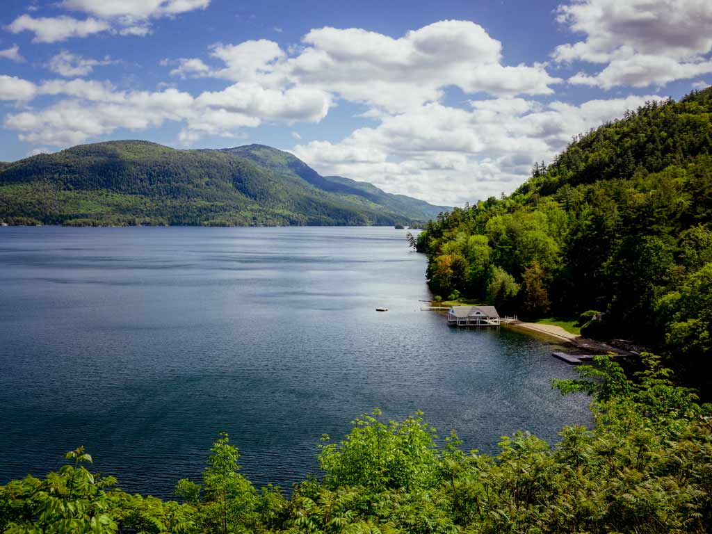 A photo of Lake George and the greenery along its shores, with forest hills in the background and a small boat house visible on the right of the image on a sunny day.