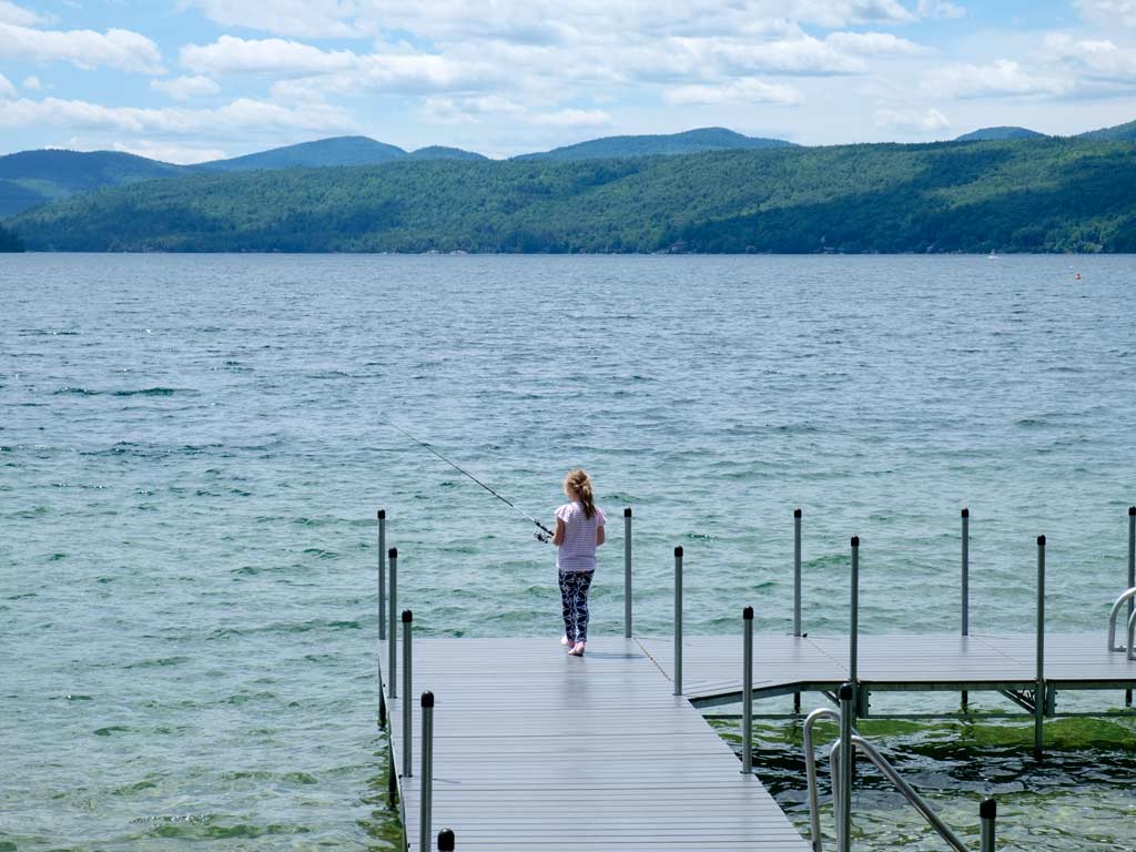 A view from a hill of a young girl fishing from a dock on Lake George on a sunny day with choppy waters.