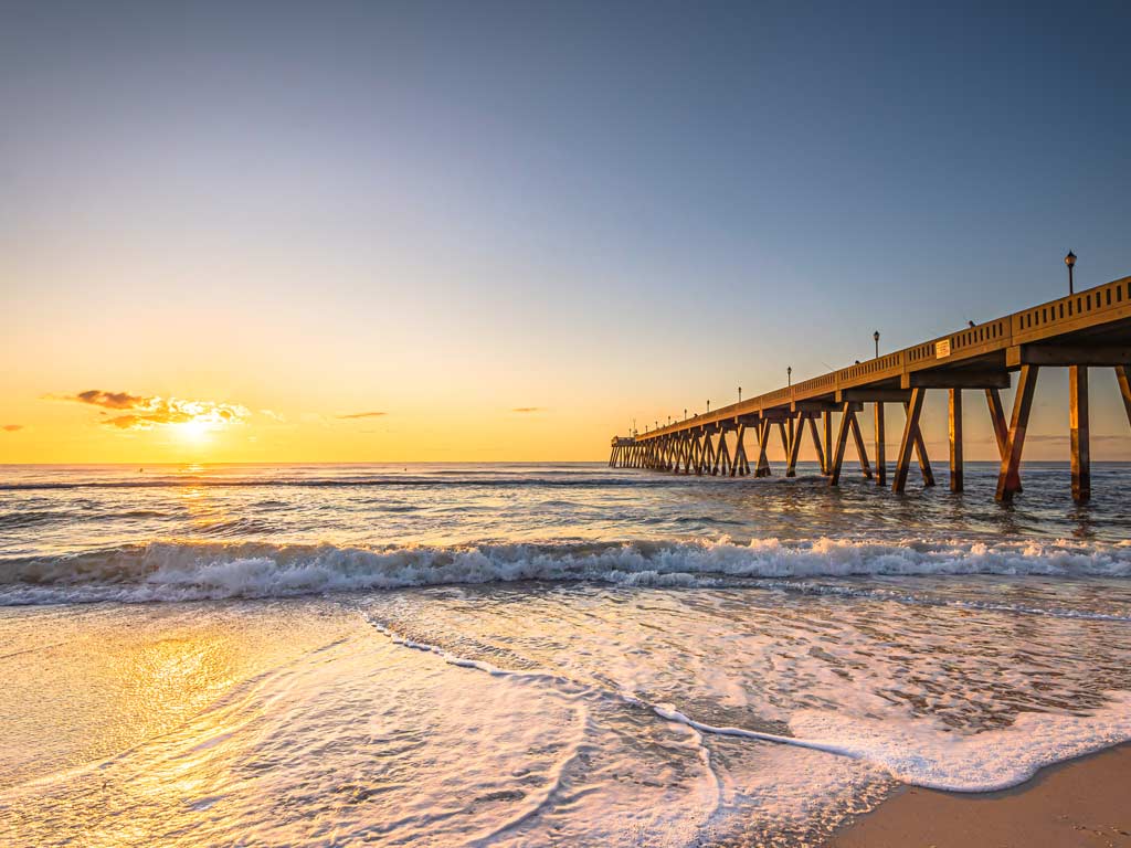 A sunset photo of a fishing pier on North Carolina's Atlantic coast, as taken from the beach with the sun setting in the distance.