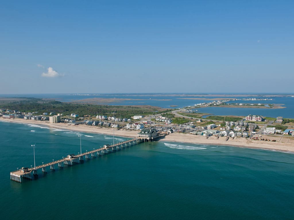 An aerial photo of the Outer Banks near Nags Head in North Carolina, with a fishing pier sticking out ionto the ocean in the foreground and a sound visible in the distance beyond the barrier island.