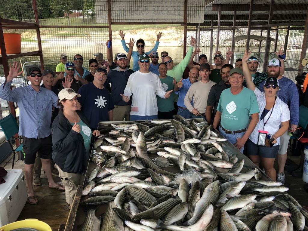 An amazing group photo of more than two dozen happy anglers posing on the dock behind the table full of fish they caught during a fishing trip earlier that day