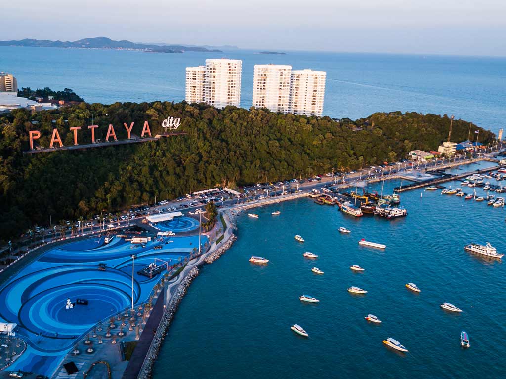 An aerial photo of the harbor in Pattaya, with different boats in the water, several large pools visible, and with a sign saying "Pattaya City" on a hill overlooking the sea. 