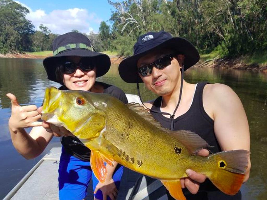 Two anglers wearing sunglasses and hats, standing on a freshwater fishing charter, holding a sizeable Peacock Bass, with the water and shoreline behind them on a sunny day