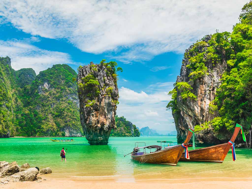 A photo of two beached Thai boats with rock formations and a man visible in the azure waters behind them on a sunny day.