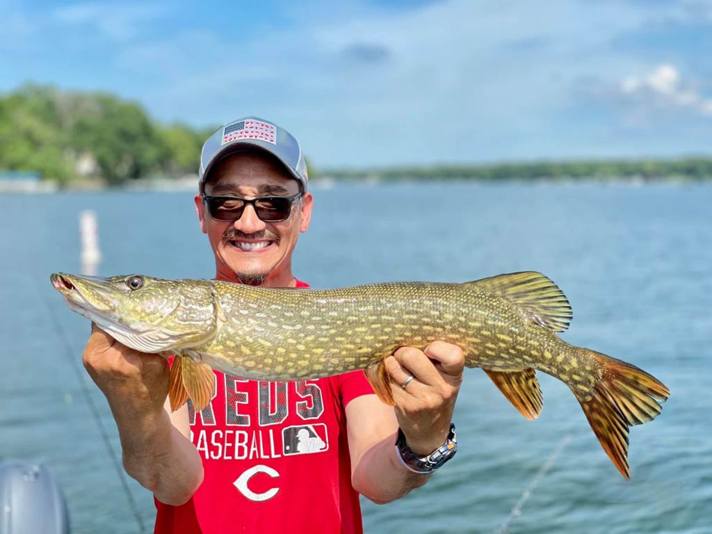 A photo of a happy angler holding a Pike with both hands while standing on a charter fishing boat during a bright and sunny day and posing against the shoreline in the background