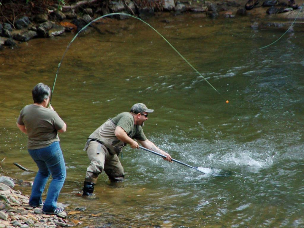 An excellent side shot of two anglers engaged in shore fishing action where one angler is standing on the river bank with a fly fishing rod and another is in the water with a net, trying to net a fish on the end of the fishing line