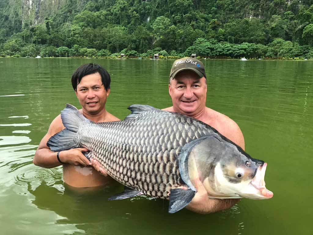 Two shirtless anglers waist deep in green waters, holding a big Siamese Carp they caught while resort fishing in Thailand.