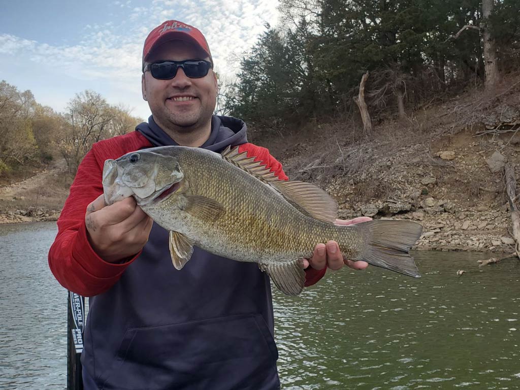 A photo of a smiling angler wearing a cap and a pair of sunglasses while holding a Smallmouth Bass with both hands and posing on a charter fishing boat against a background of trees and the shoreline 