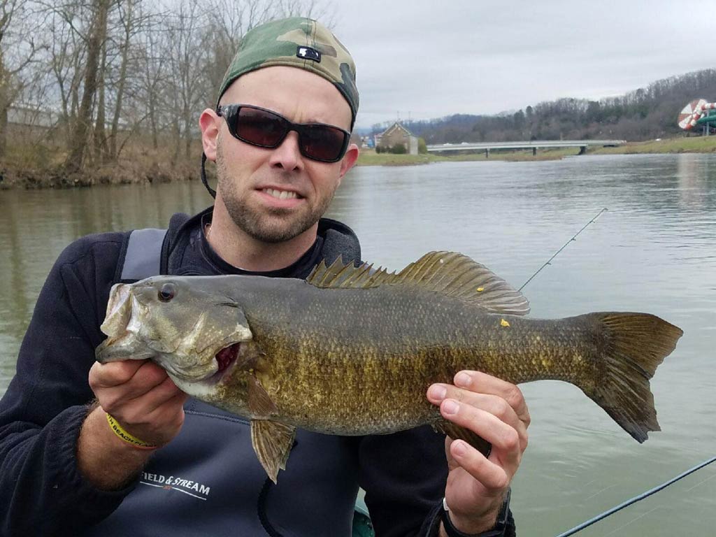 A photo of an angler wearing a cap and sunglasses while sitting on a charter fishing boat and holding a big Smallmouth Bass with both hands, caught on the Tennessee River during the fall season