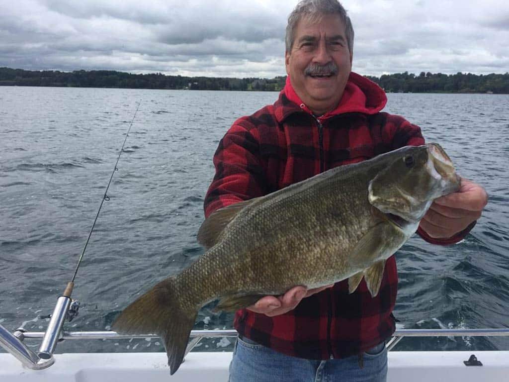 A smiling angler in a red jacket on a boat, holding a sizeable Smallmouth Bass, with a fishing rod trolling off the side of the boat to his right on a cloudy day.