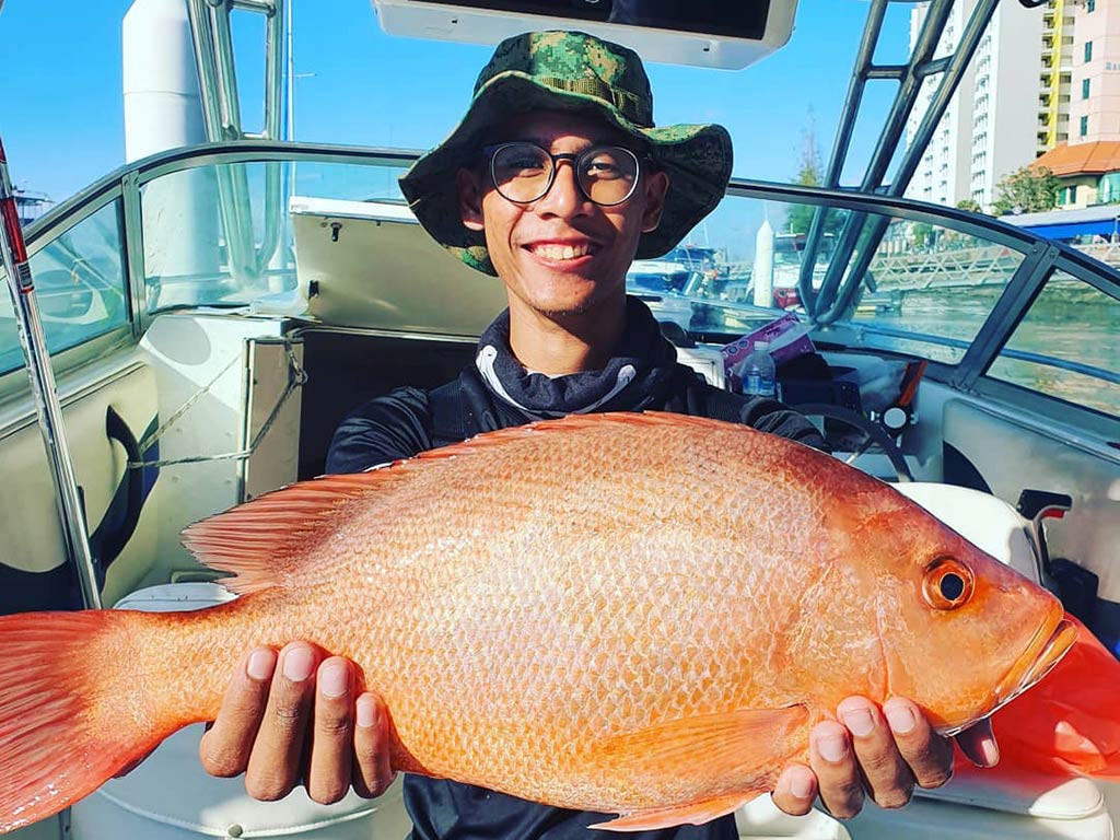 A smiling angler in a bucket hat holds a large Red Emperor Snapper aboard a fishing boat, with the boat's bridge visible behind him