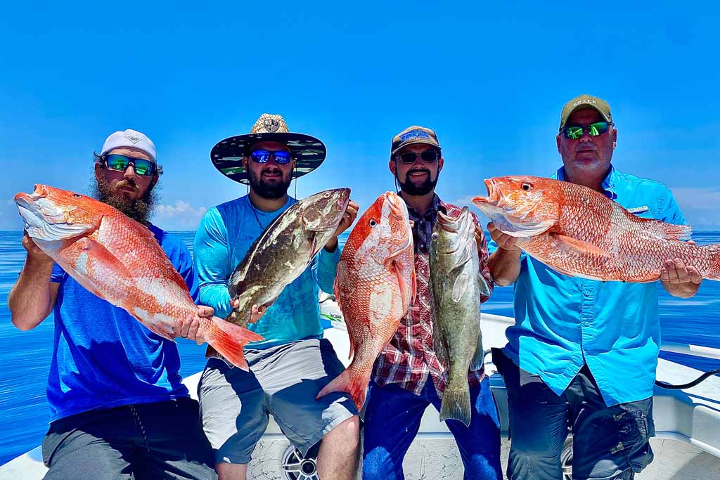 Four fishermen standing on a charter fishing boat, each holding a Snapper and/or Grouper, with blue skies and open waters in the background