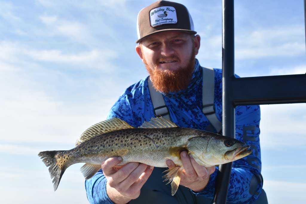 A smiling man in a cap and sporting a ginger beard holding a Spotted Seatrout close to the camera on a sunny day