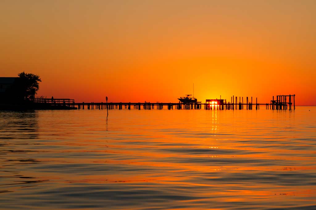 A view across the water of the St. George Island Pier at sunset, with the pier as a silhouette against the orange-red sky