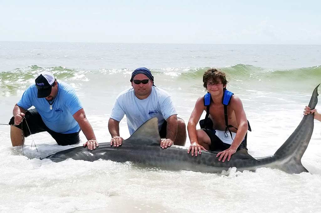 Three anglers standing in the surf at St. George Island, holding a big Shark on the ground as the waves crash in behind them