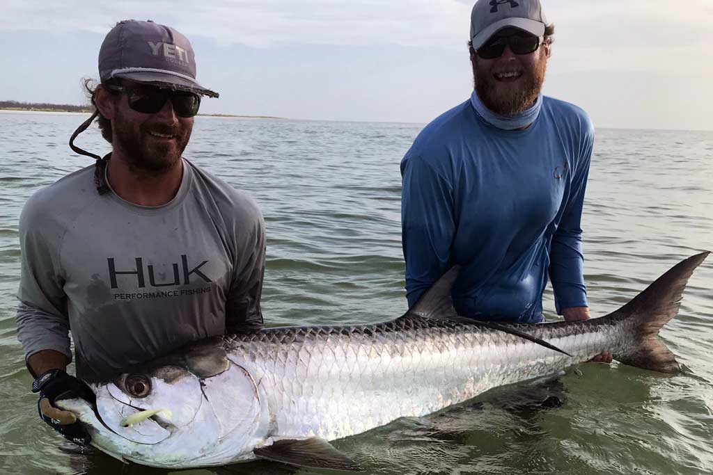 Two smiling fishermen standing in water up to their waist, holding a Tarpon, half-submerged in water