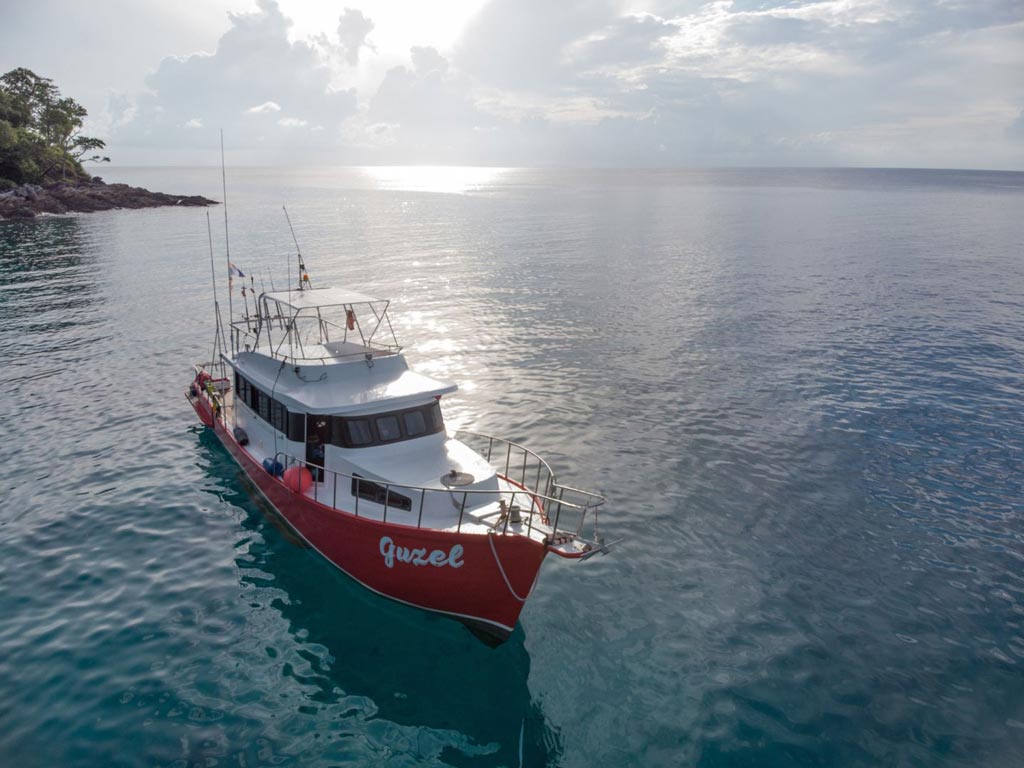 An aerial photo of a red and white offshore fishing boat in the sea, with its name, "Guzel" written on its hull and the sun reflecting off the water surface.