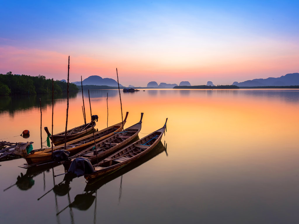 A view of three Thai longboats in the water at sunset, with their shadows reflecting off the calm water surface.