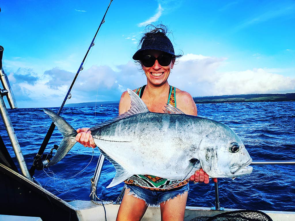 A female angler standing on a boat and smiling while holding a large Trevally, with the water behind her on a sunny day