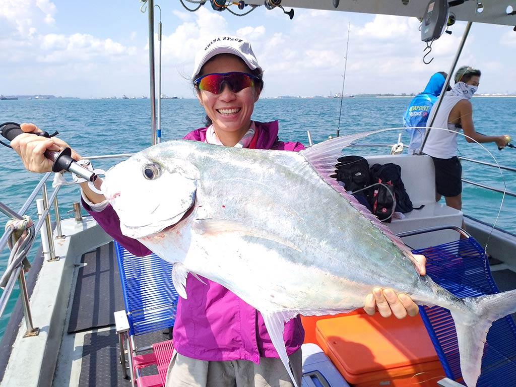 A female angler in a cap and sunglasses holding a large Diamond Trevally on a fishing charter in Singapore