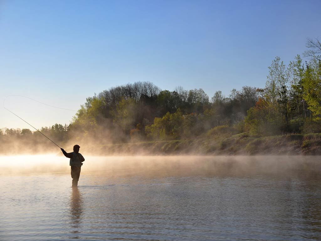 A photo of an angler holding a fly fishing rod in one hand preparing to cast while standing in the water facing the shoreline and wading on an early fall morning while there’s still fog above the water’s surface