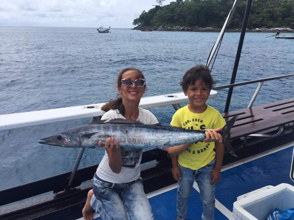 A female angler kneeling on the deck of a fishing boat in Thailand, holding a Wahoo she caught with a smiling child posing next to her.