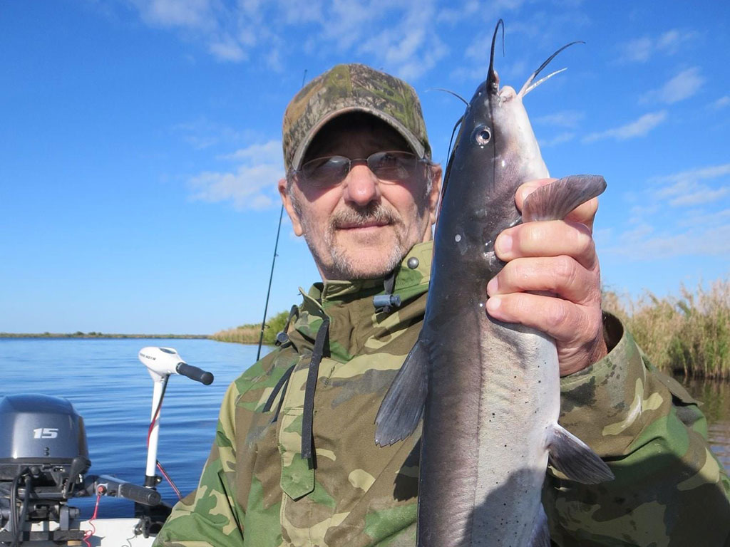 A closeup of a man in a cap sitting on a fishing boat in Texas and holding a Catfish upright, with the water behind him on a clear day