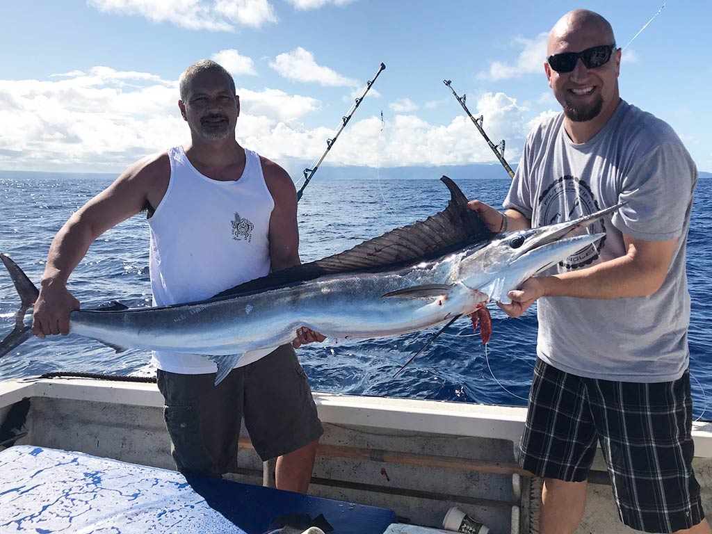 Two anglers standing on a fishing charter in Hawaii and holding a large Marlin, with water behind them and two trolling rods off the side of the boat