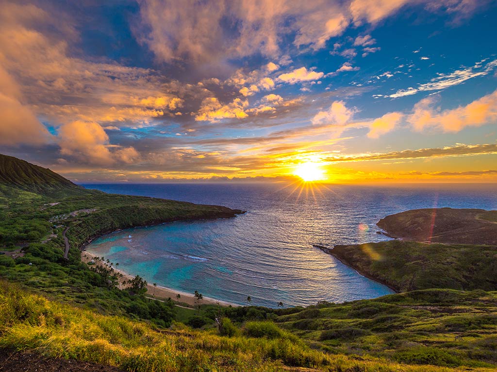 A view of a small bay in Oahu taken from a hill at sunset, with the sun in the distance on the horizon