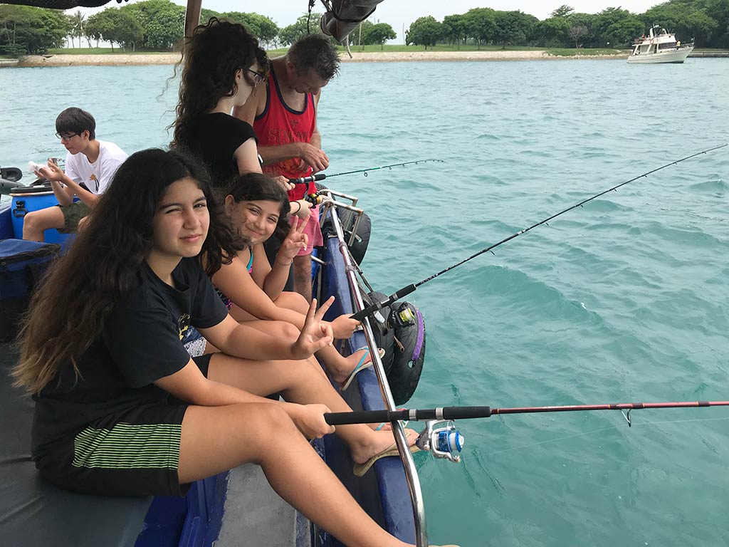 A group of young anglers sitting on the side of the boat with their rods in the water, while looking at the camera