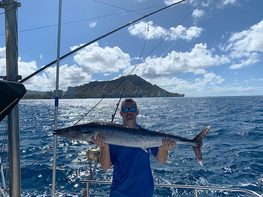 An angler standing on a boat with the water and a spit of land behind him in the distance, while holding a Wahoo caught in Oahu