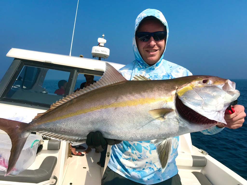 An angler wearing sunglasses and a hoodie, holding a big Amberjack reeled in while fishing in Dubai, with the center console of the boat he's on visible behind him.