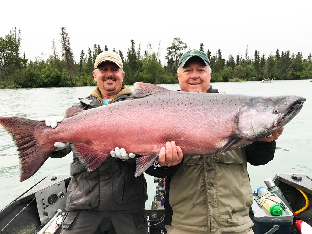 Two smiling anglers standing on a fishing boat, holding a huge Salmon, with a river and greenery in the background on a cloudy day