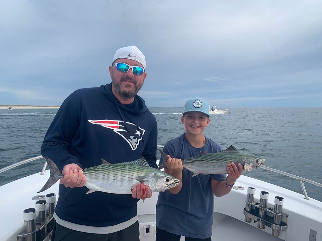 A father and son, both wearing baseball caps, standing on the front of a fishing boat, holding a Bonito each, with the waters behind them on a cloudy day