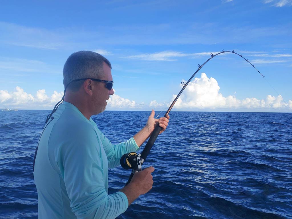 A side view of an angler standing on a charter fishing boat and holding a bended rod while bottom fishing for Snappers and Groupers 