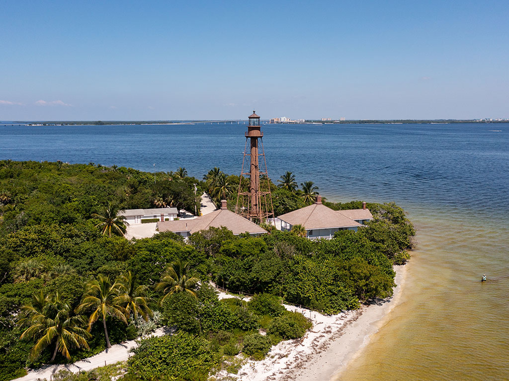 An aerial view of the lighthouse near Sanibel Island Fishing Pier, with the waters of the Gulf of Mexico on the right of the image and in the distance behind the lighthouse and some greenery