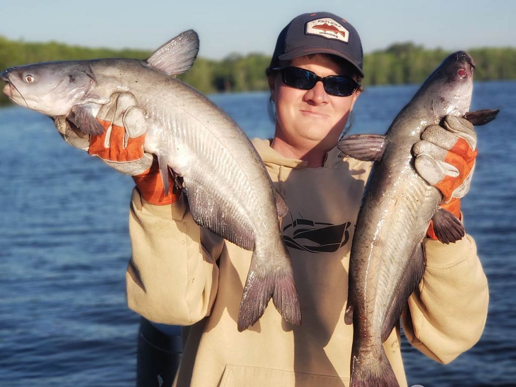 An angler in a baseball cap and sunglasses holding up two Catfish while standing on a boat with the water behind him on a sunny day