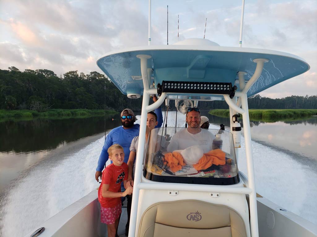 A photo featuring a group of anglers of all ages standing on a St. Simons Island fishing charter boat and posing with their captain while the captain is controlling the boat and taking them to the angling hotspot for the day