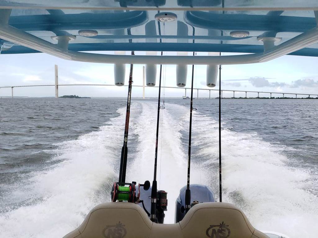 A photo of a charter fishing boat taken while fast-paced trolling, featuring four angling rods and two motors at the back of it and a white bridge in the distance