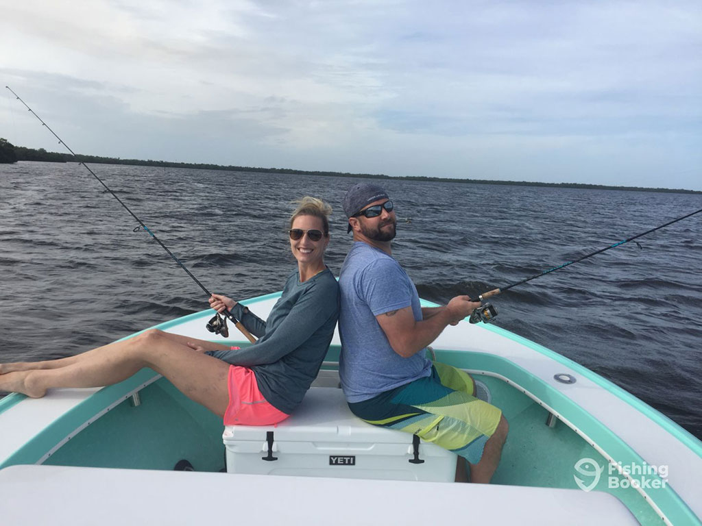 A male and female angler sit back to back while holding a fishing rod each on a fishing charter, with the water in the distance