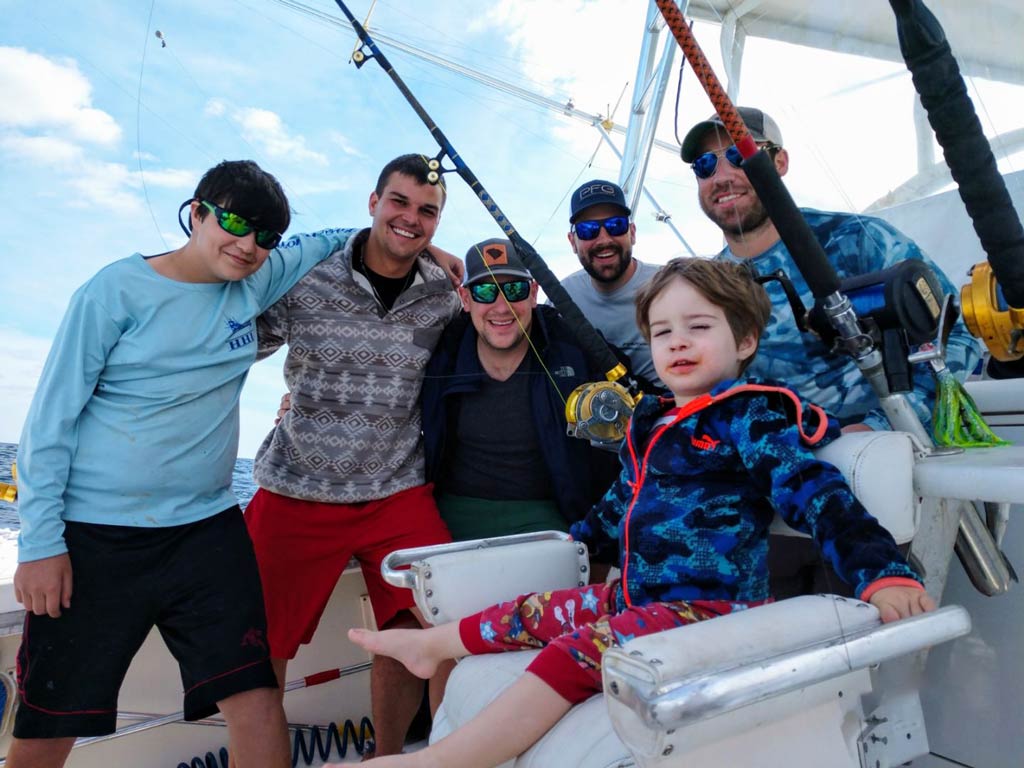 A group photo of several anglers standing, while a kid sits on a fighting chair – all posing with their captain on a charter fishing boat during their deep sea fishing trip