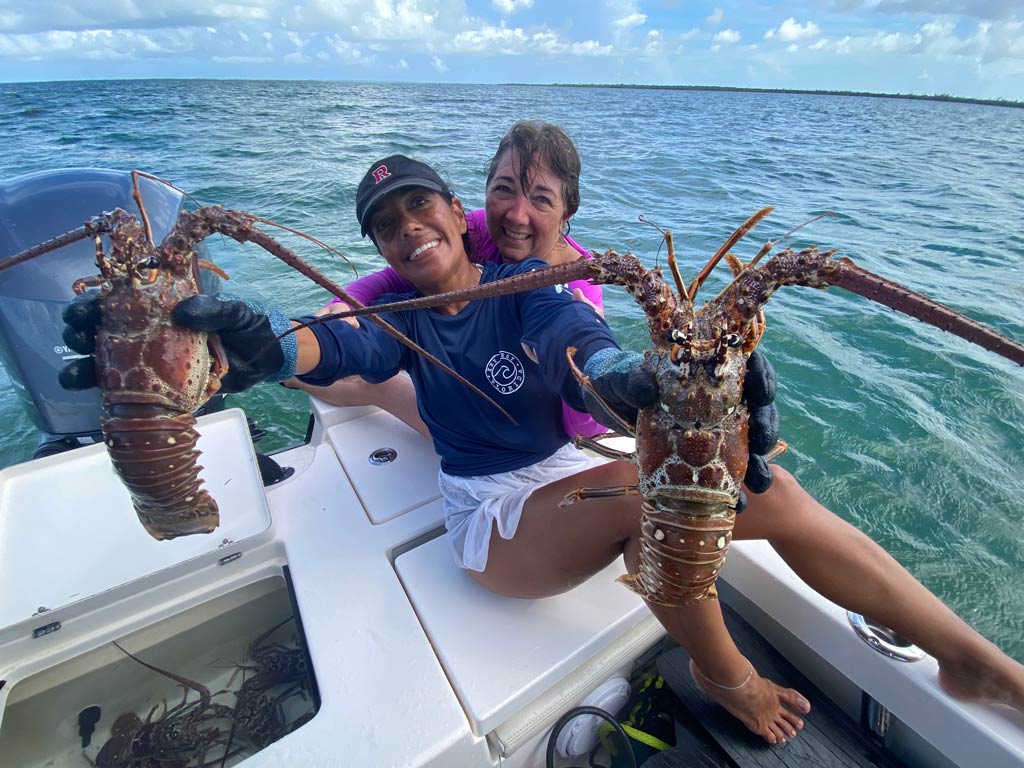 A photo of two female anglers smiling and sitting on a charter boat while one of them is wearing gloves and happily posing for the camera with a Lobster in each hand