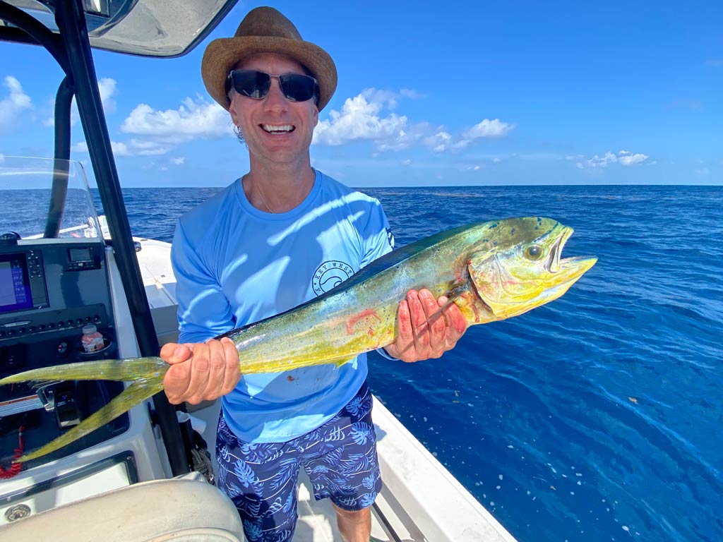 A photo of an angler wearing a hat and a pair of sunglasses while standing on a Cudjoe Key fishing charter boat and holding small Mahi Mahi caught in the offshore waters on a bright summer day