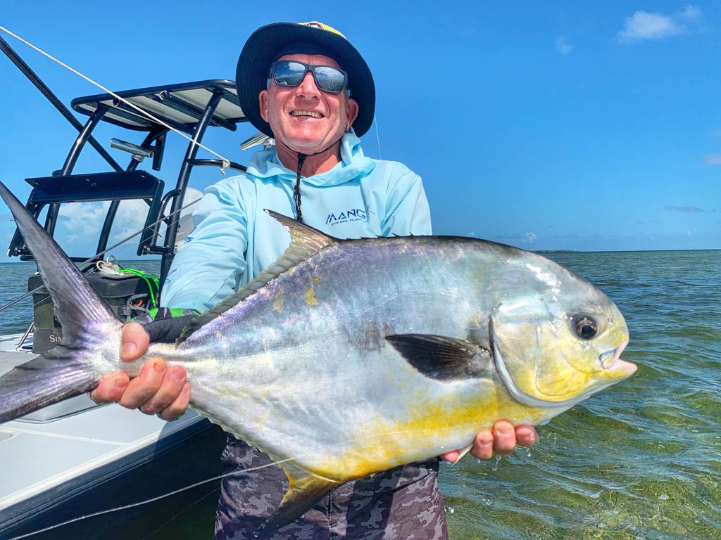 A photo of a proud angler wearing a hat and a pair of sunglasses while posing with a big Permit fish with his Cudjoe Key fishing charter in the background