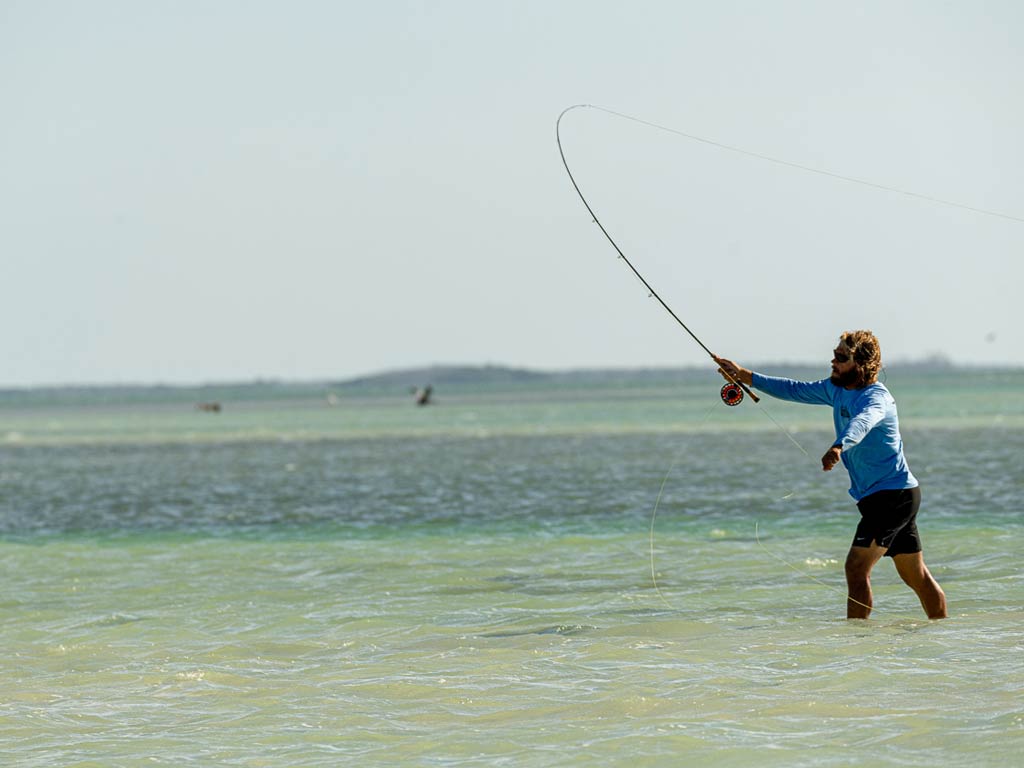 A photo of an angler standing in the ankle-deep water and casting a fly fishing rod in the shallows on a bright and sunny day