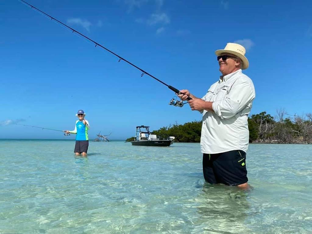 A photo of two happy anglers posing while standing in the water and fly fishing with their boat standing still in the background on a bright and sunny day